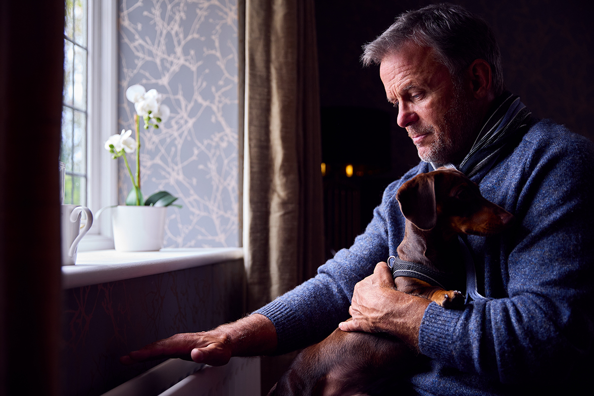 Mature-Man-With-Pet-Dog-Trying-To-Keep-Warm-By-Radiator-At-Home-During-Cost-Of-Living-Energy-Crisis
